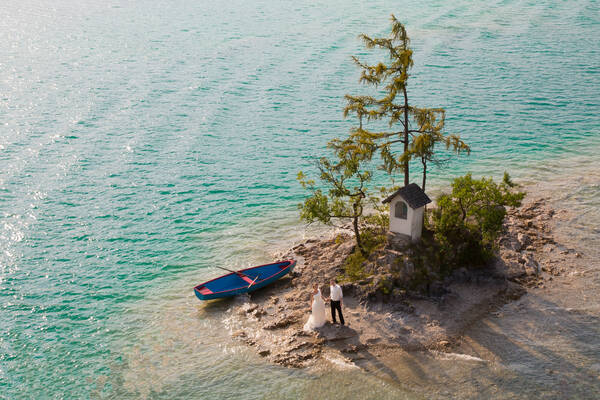 Ein Brautpaar aus der Luft Fotografiert auf einer winig kleinen Insel in einem See mit Bildstock. Neben dem Paar ist ein blaues Boot mit roten Rudern.