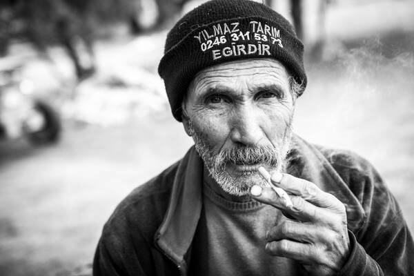 Ein alter bärtiger Mann trägt eine dunkle Mütze mit Aufschrift (Name, Telefonnummer und der türkische Ortsname Egirdir) und raucht eine Zigarette. Schwarz-Weiß.