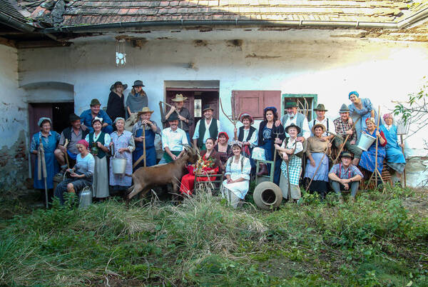 Gruppenbild von bäuerlich gekleideten Menschen vor einem Bauernhof. Viele haben alte Handwerks- und Arbeitsgegenstände dabei. Im Vordergrund sind auch zwei Ziegen mit im Bild, die gerade versuchen, aus einem Obstkorb zu Fressen, den ein rot gekleidetes Mädchen am Schoß hat..