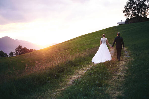 Ein Brautpaar geht händehaltend bei Sonnenuntergang einen Fahrweg durch eine Wiese bergan.
