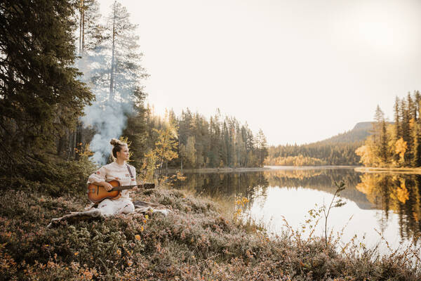 Eine junge, hell gekleidete Frau sithe auf einer Decke am Ufer eines herbstlichen Bergsees und spielt Gitarre. Hinter ihr steigt Rauch wie von einem Lagerfeuer auf.