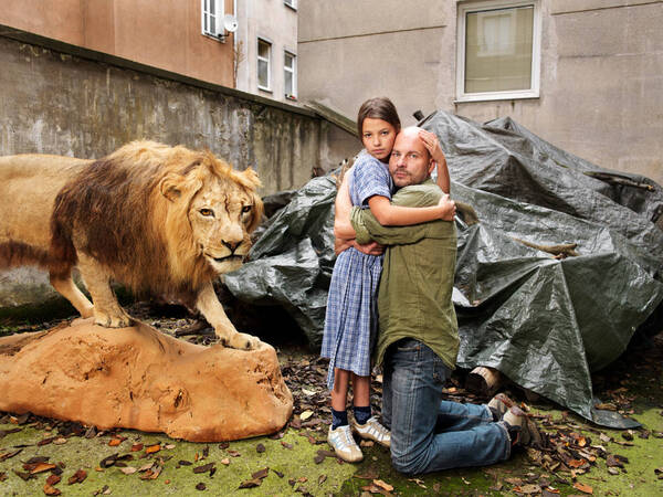 Ein kniender Mann wird von einem Mädchen in einem Hinterhof umarmt. Ein Ausgestopfter Löwe steht hinter ihnen.