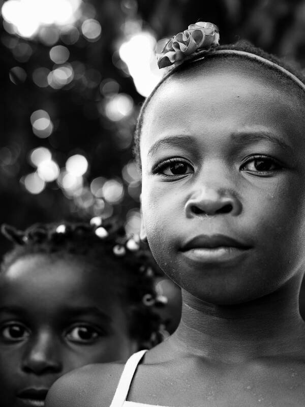 ein dunkelhäutiges Mädchen mit einem Haarreifen mit Stoffblume blickt in die Kamera. ein weiteres dunkelhäutiges Mädchen schaut ihm über die Schulter.