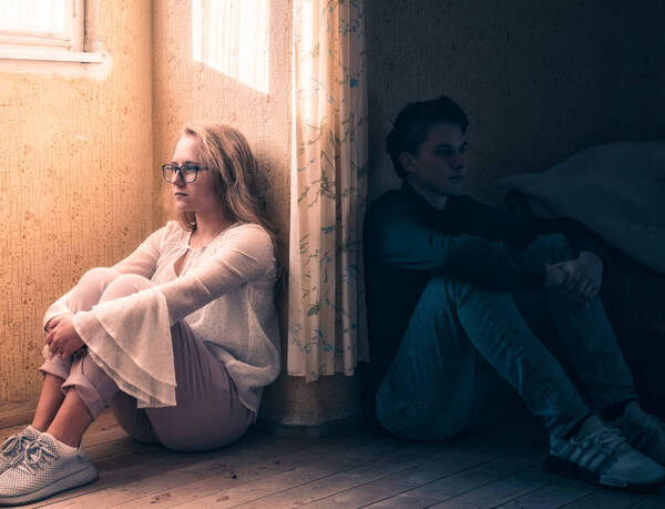 Ein Mann und eine Frau sitzen mit dem Rücken an verschiedenen Wänden eines Zimmers und blicken in unterschiedliche Richtungen.