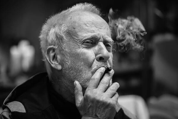 Nahaufnahme eines weißhaarigen, bärtigen Mannes, der gerade an einer Zigarre zieht. Schwarz-Weiß