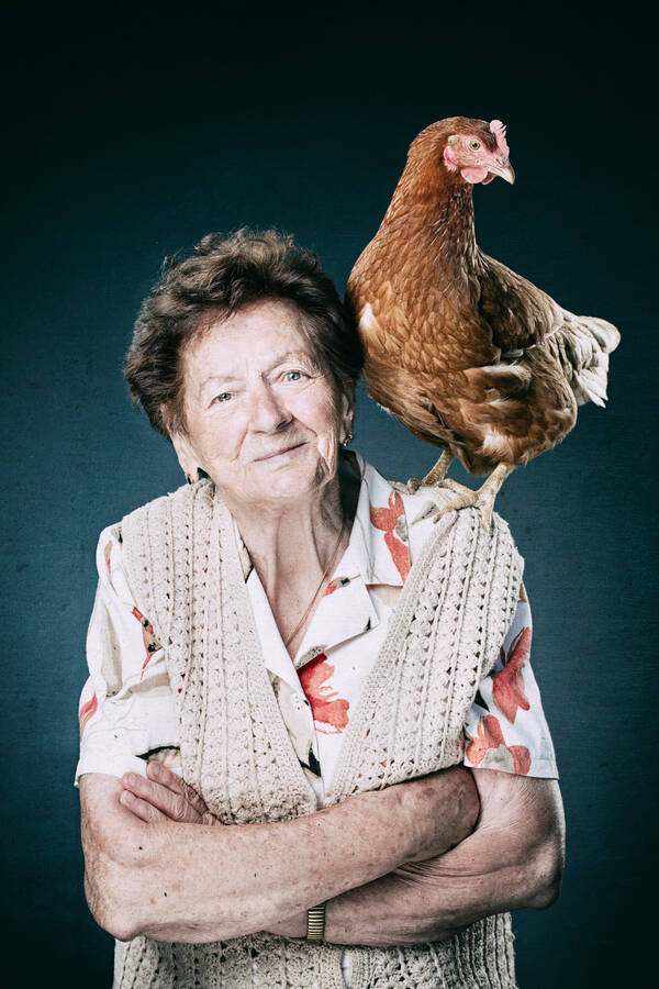 Eine alte Frau in einem weiß-roten Blumenkleid und beiger Strickjacke steht mit verschränkten Armen. Auf ihrer linken Schulter steht ein braunes Huhn.