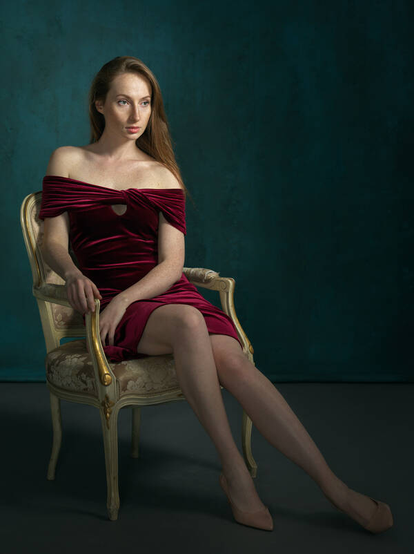 Eine Langhaarige Frau sitzt mit einem schulterfreien dunkelroten Samtkleid in einem barocken Sessel.