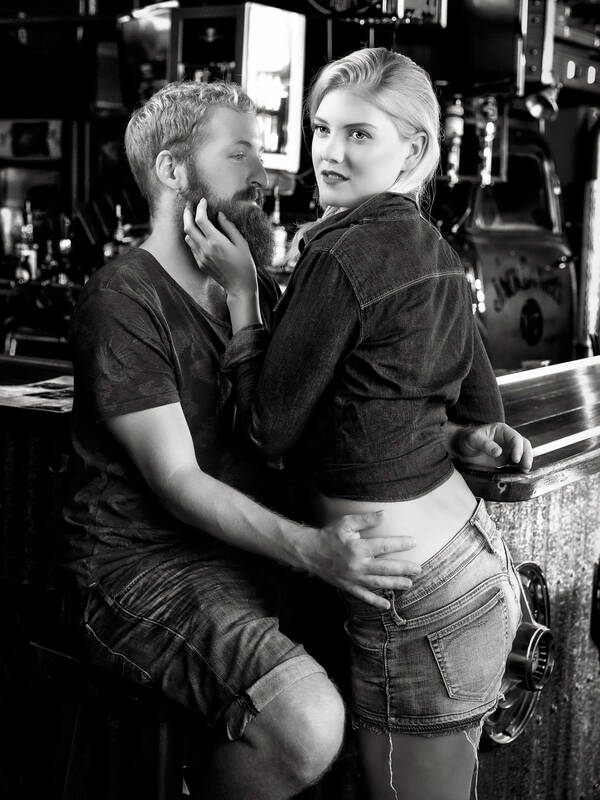 Ein Paar an einer Theke, der bärtige Mann sitzt auf einem Barhocker und schaut auf die Frau, die er  um die Hüfte hält. Die Frau in fransigen Hotpants legt dem Mann die Hand ans Kinn und schaut in eine andere Richtung. Schwarz-Weiß.