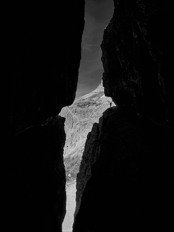 Blick durch einen Spalt in Felsen: auf einem Felsband hebt sich die winzige Silhouette eines Bergsteigers vor den Felswänden im Hintergrund ab. Schwarz-Weiß