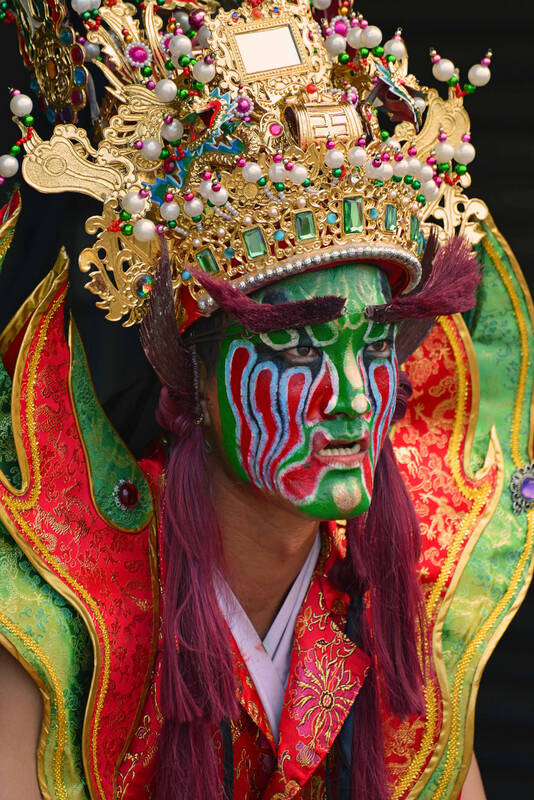 Ein Mann mit geschminktem Gesicht, fernöstlicher Krone und grün-rotem Kostüm.