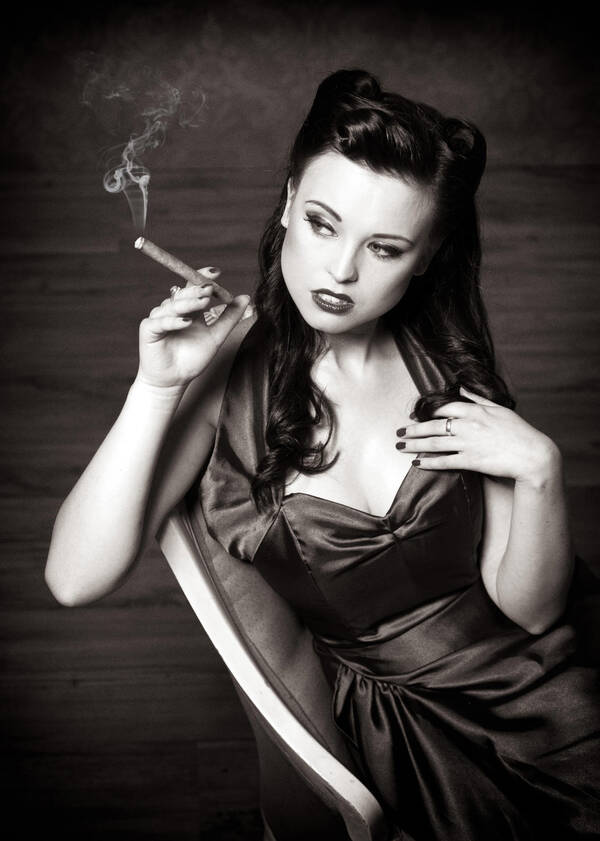 Eine Frau in elegantem, dunklem Seidenkleid lehnt auf einem Stuhl und raucht eine Zigarre. Schwarz-Weiß.