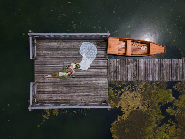 Ein Brautpaar (er in Tracht, sie in weißem Kleid mit Schleppe) liegt auf einem Landungssteg neben einem Ruderboot im See.