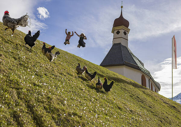 Zwei Kinder springen neben einer Kapelle  in einen steilen Wiesenabhang voller Krokusse. Im Vordergund ist ein Hahn mit sieben Hennen gerade im Begriff zu flüchten.