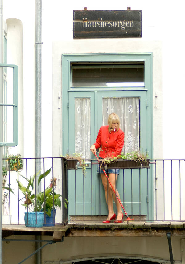 Auf einem Balkon vor einer Türe kehrt eine blonde Frau in roten Highheels, Jeans-Minirock und roter Jacke mit einem roten Besen. Im Mundwinkel hat sie lässig eine Zigarette. Über der Türe hinter ihr hängt ein Schild mit der Aufschrift: Hausbesorger.