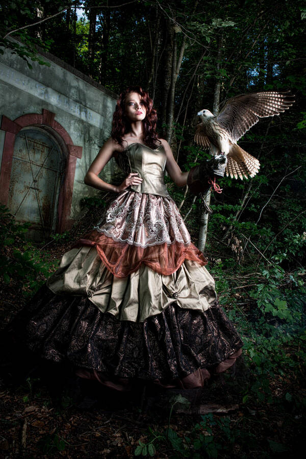 Eine junge Frau steht in einem weit ausladenden, vielschichtigen Kleid vor einem Portal im Wald. Auf der Hand hat sie einen abgerichteten Greifvogel.