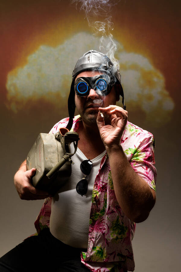 Ein Mann mit Kanister im Arm, altem Fliegerhelm und -brille in Hawaihemd raucht neben dem offenen Kanister eine Zigarette. im Hintergrund ein Atompilz.