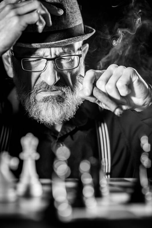 Ein alter bärtiger Mann mit Hut und Brille  sitzt Zigarre-rauchend vor einem Schachspiel. Schwarz-Weiß.