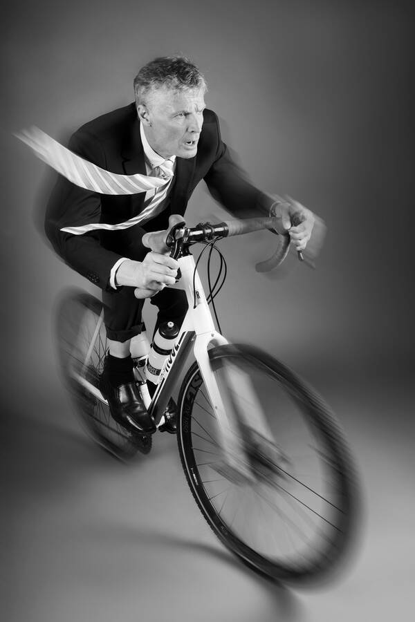 Ein Mann in dunklem Anzug fährt schnell mit einem Mountain-Bike. Beim rechten Bein ist die Hose aufgekrempelt. Die gestreifte Krawatte fliegt im Wind. Bewegungsunschärfe im vorderen und hinteren Bereich des Rades. Schwarz-Weiß.