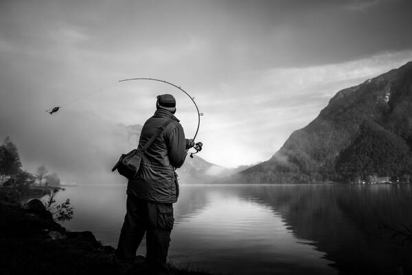Ein Angler steht vor einem See, die Rute in seiner Hand krümmt sich in der Bewegung. An der Leine hängt ein kleiner Fisch. Schwarz-Weiß.