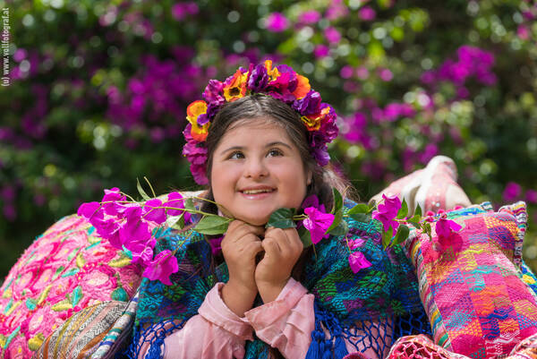 Eine junge Frau umgeben von bunten Stoffen mit einem Blumenkranz. Verträumter Blick.
