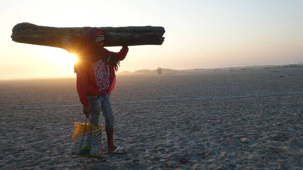 Ein Mann in Sandalen mit einem Kopftuch um den Kopf geschlungen trägt bei Sonnenaufgang ein großes Stück Treibholz und eine Tasche durch den Sand.
