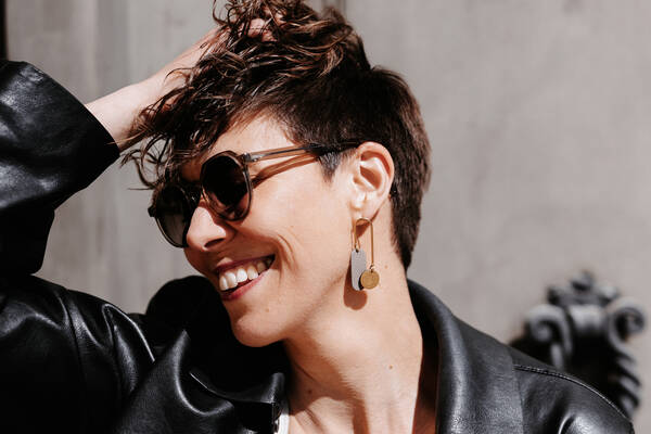 Portrait einer kurzhaarigen Frau mit Sonnenbrille und Lederjacke, die sich mit der rechten Hand in die Haare greift.