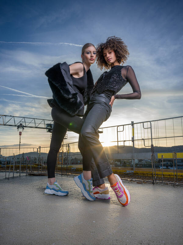 Zwei junge Frauen im Abendlicht in schwarzer jugendlicher Kleidung mit poppigen Sneakers fotografiert auf einer frisch betonierten Deckenplatte einer Baustelle.