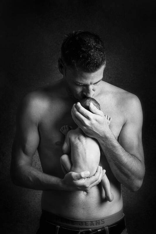 Ein Mann mit nacktem Oberkörper hält ein neugeborenes Kind vor seiner Brust und küsst es auf den Kopf. Schwarz-Weiß