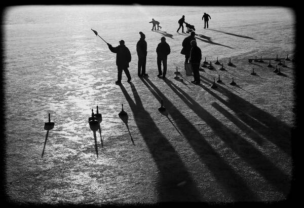 Fünf Männer auf einer zugefrorenen Wasserfläche beim Eisstockschießen. Die tiefstehende Sonne lässt von den Männern und Eisstöcken nur die Silhouetten erkennen und wirft lange Schatten. Im Hintergrund sieht man Menschen beim Eislaufen. Schwarz-Weiß.