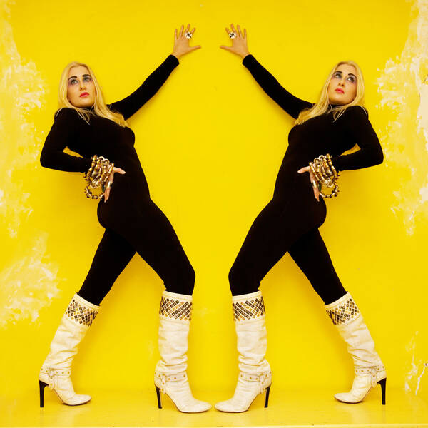 Eine blonde, schwarz gekleidete Frau mit hohen weißen Stiefeln und  üppigem Schmuck vor einer gelben Wand - gespiegelt.