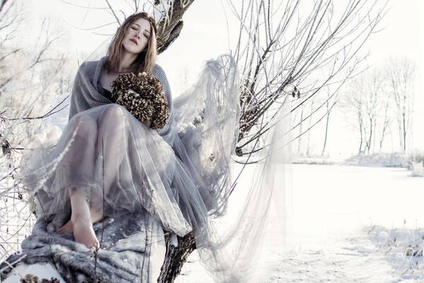 Eine junge Frau in Schleier gehüllt sitzt in winterlicher Landschaft auf einem Baum