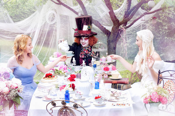 Zwei Frauen sitzen mit dem mad Hatter bei einer Tee-Jause im Garten.