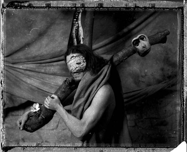 Portrait eines langhaarigen Mannes, dessen Nackter Oberkörper nur von einem Tuch verhüllt ist. Der Mann Tträgt einen Baumstamm über der Schulter, auf dem eine Affenmaske hängt. Über dem Gesicht trägt der Mann selbst auch eine Maske die wie Baumrinde oder Krokodilleder strukturiert ist. Auf dem Kopf trägt er eine Krokodilschnauze. Ein zweiter Baumstamm hinter dem Mann erzeugt gemeinsam mit dem ersten Baumstamm den eindruck, als ob der Mann wie Jesus Christus ein Kreuz tragen würde. Im Hintergrund sind Tücher über eine Steinmauer gehängt. Das Bild zeigt an den Rändern den typischen Rand von analogen Negativen und ist voller Staub- und anderer Verunreinigungen. Schwarz-Weiß.