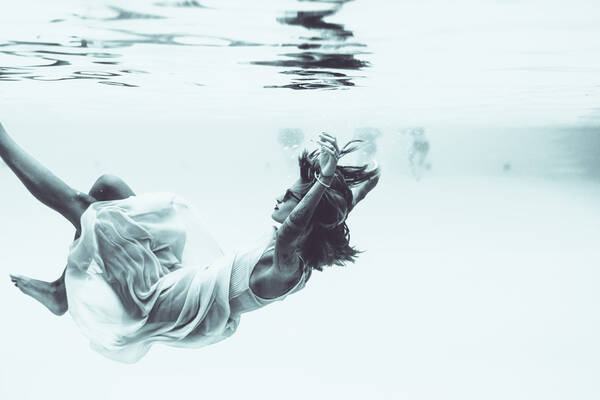 Eine junge Frau treibt mit Gewand und Sonnenbrille durch das Wasser eines Pools. sie spiegelt sich in der Wasseroberfläche ober ihr. Im Hintergrund sieht man Schwimmer.