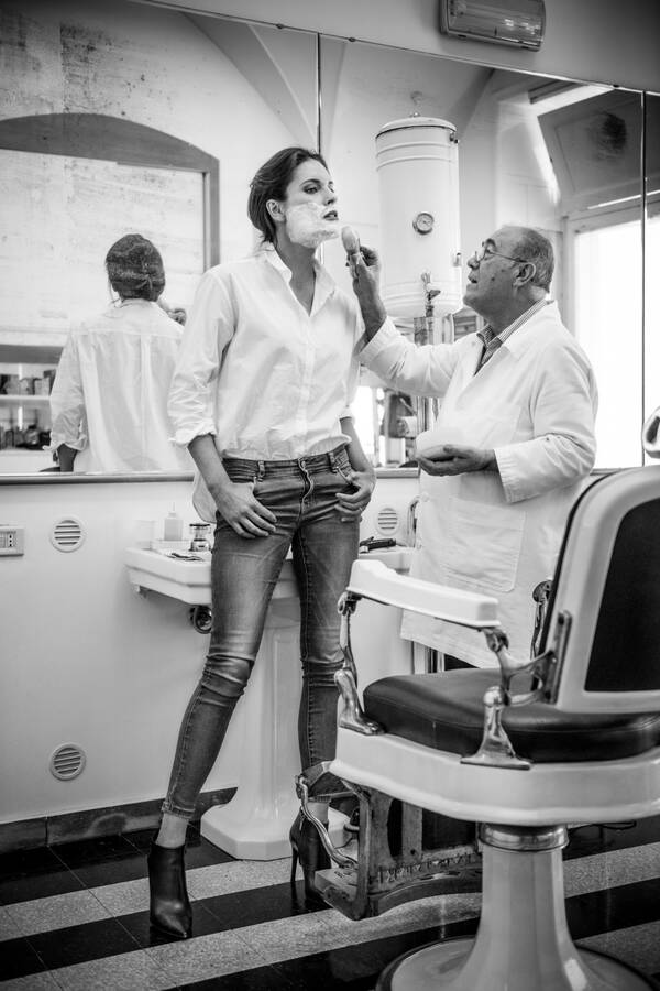 Ein älterer untersetzter Barbier/Frieseur schäumt einer stehenden großgewachsenen Frau das Gesicht ein wie zum Rasieren. Schwarz-Weiß.