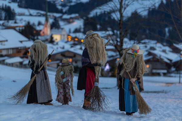 Hexenfiguren einer Brauchtumsgruppe vor einem winterlich verschneitem Dorf in der Dämmerung.