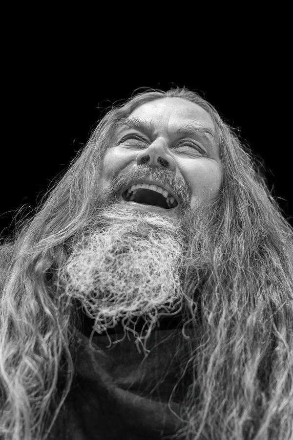Portrait eines lachenden, bärtigen, langhaarigen Mannes vor schwarzem Hintergrund. Schwarz-Weiß