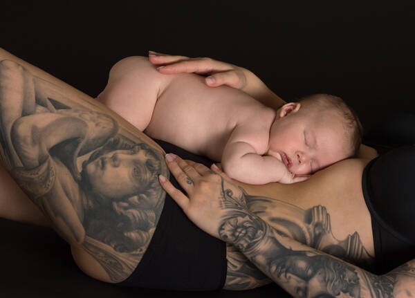Ein Baby liegt am Bauch einer stark tätowierten Frau in schwarzer Unterwäsche.