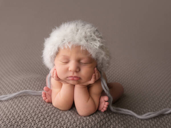 Neugeborenes Baby mit flauschiger, weißer Mütze in der Frosch Pose.