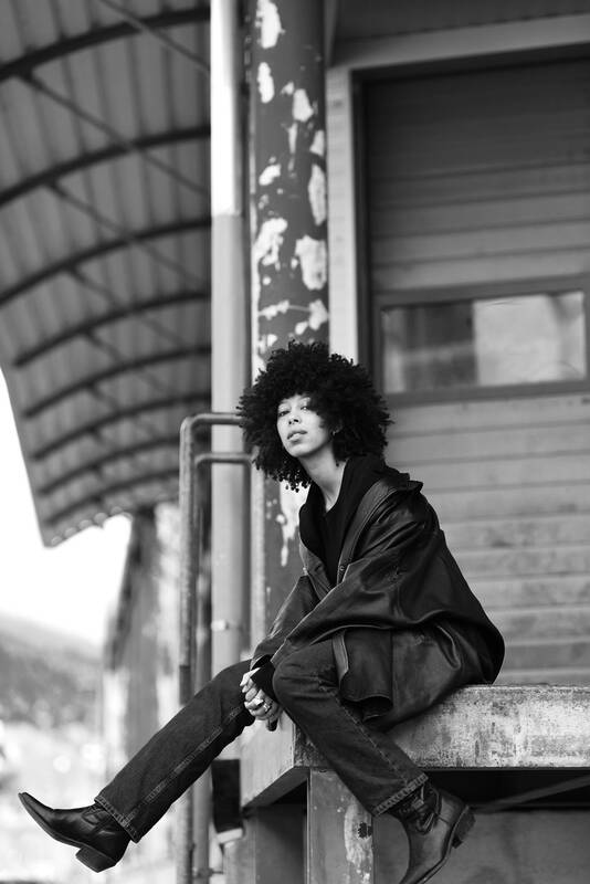 Eine junge Frau in Lederjacke und Boots mit Afro-Frisur sitzt auf einer Laderampe. Schwarz-Weiß.