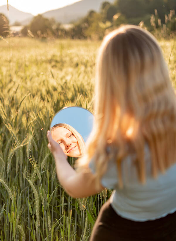 Eine junge Frau steht im Abendlich vor einem Getreidefeld und sieht sich selbst in einem runden Spiegel, den sie in der Hand hält.