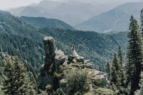 Ein Brautpaar steht auf einer felsigen Erhebung in einem Gebirgswald mit Blick auf die bewaldeten Hügel, Täler und Berge ringsum.