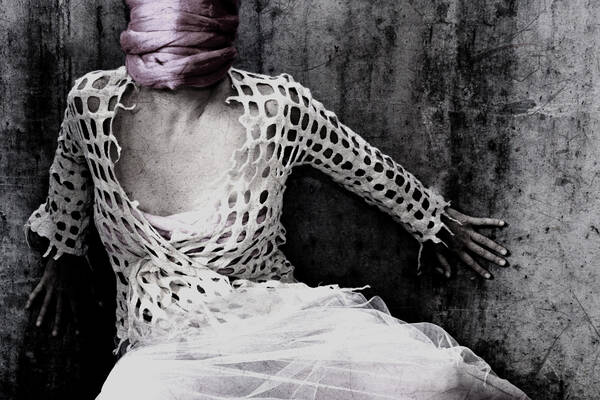 Eine Frau in weißen Kleidern mit verhülltem Gesicht, die sich angstvoll gegen eine Betonwand drückt.