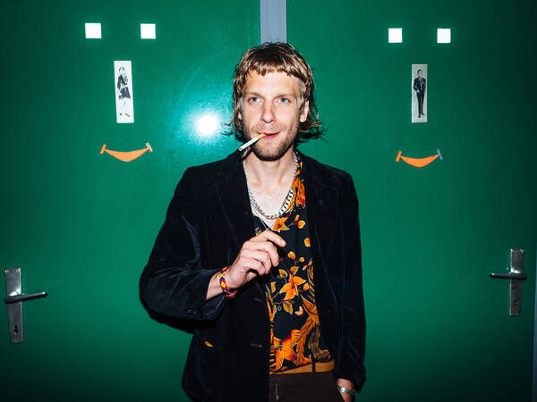 Portait von Voodoo Jürgens mit einer Zigarette im Mund vor grünen Toilettentüren.