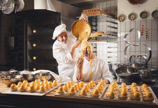 In der Backstube der Konditorei sitzt ein bärtiger Konditor mit Brille an einem Arbeitstisch auf dem mehrere Bleche mit bereits fertig verzierten Pöllauer Piperln stehen. Rund um ihn sind Edelstahlschüsseln und Arbeitsgeräte. Ein junger Mitarbeiter kippt ihm von hinten aus einer Schüssel Glasur über den Kopf.