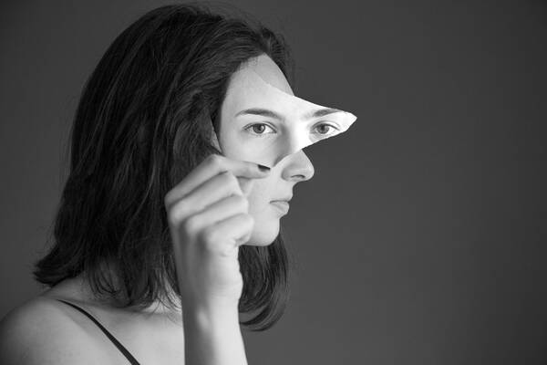 Portrait einer jungen Frau im Profil. Mit der rechten Hand hält sie sich ein Stück eines Portraits vor das Gesicht, auf dem zwei Augen zu sehen sind. Schwarz-Weiß.
