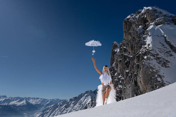 Eine in weißem Bühnenoutfit gekleidete Frau stapft durch eine winterlich verschneite Berglandschaft und wirlft einen weißen Schirm in die Luft.