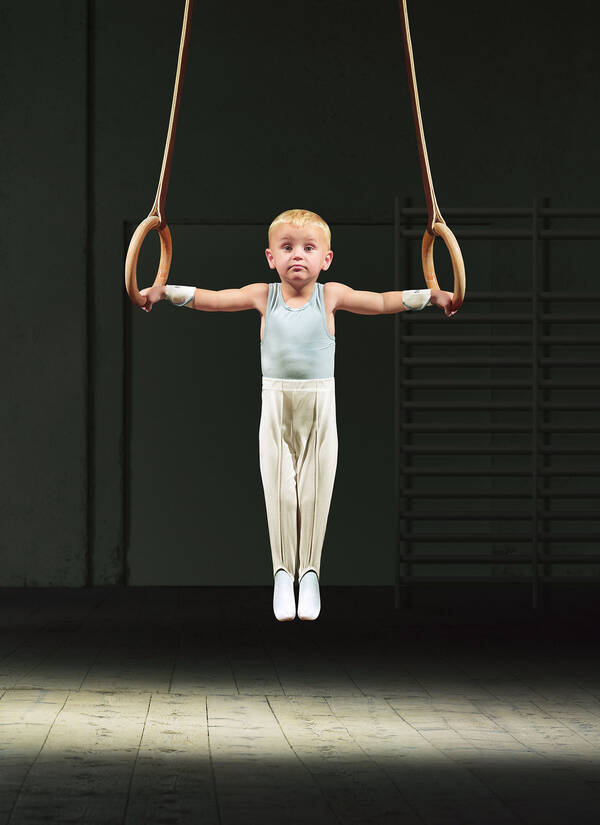 in einer dunklen Turnhalle führ in einem Lichtkegel ein kleiner, blonder Bub im Athletik-Trikot scheinbar mühelos einen Kreuzhang an den Ringen aus.