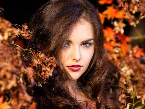 Portrait einer jungen, dunkelhaarigen Frau mit dunkelrot geschminkten Lippen zwischen Herbstlich rot-orange gefärbten Bllättern.