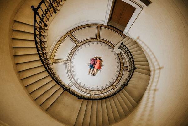 Blick von oben in ein Stiegenhaus, das sich schneckenhausartig nach unten schraubt. Auf der untersten Ebene liegt ein Paar auf dem Fußboden.
